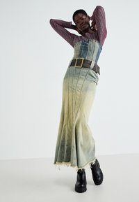 Robe maxi en denim dégradé de bleu clair à vert, avec un corsage structuré et une large ceinture marron. Bottes à plateforme noires avec semelles texturées.
