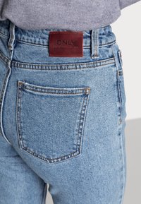 Blå denimjeans med en brun läderpatch med prägling, bakficka med sömnadsdetaljer och en klassisk hög midja.
