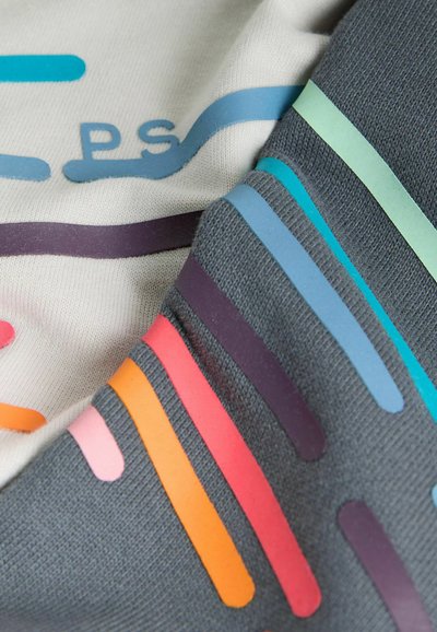 Nærbillede af gråt og hvidt stof med farverige horisontale striber og delvist tekst "PS" i blåt på det hvide stof.