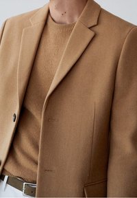 Blazer de mezcla de lana marrón con solapas en pico, dos botones y bolsillos laterales, superpuesto a un suéter marrón a juego, que presenta una textura acanalada.