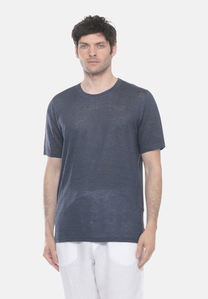 Uomo con capelli ricci scuri che indossa una maglietta blu navy a maniche corte e pantaloni bianchi, in piedi contro uno sfondo chiaro e semplice.