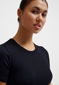 Young woman with dark hair tied back wearing small gold hoop earrings, a thin gold necklace, and a ribbed black short-sleeve top against white background.