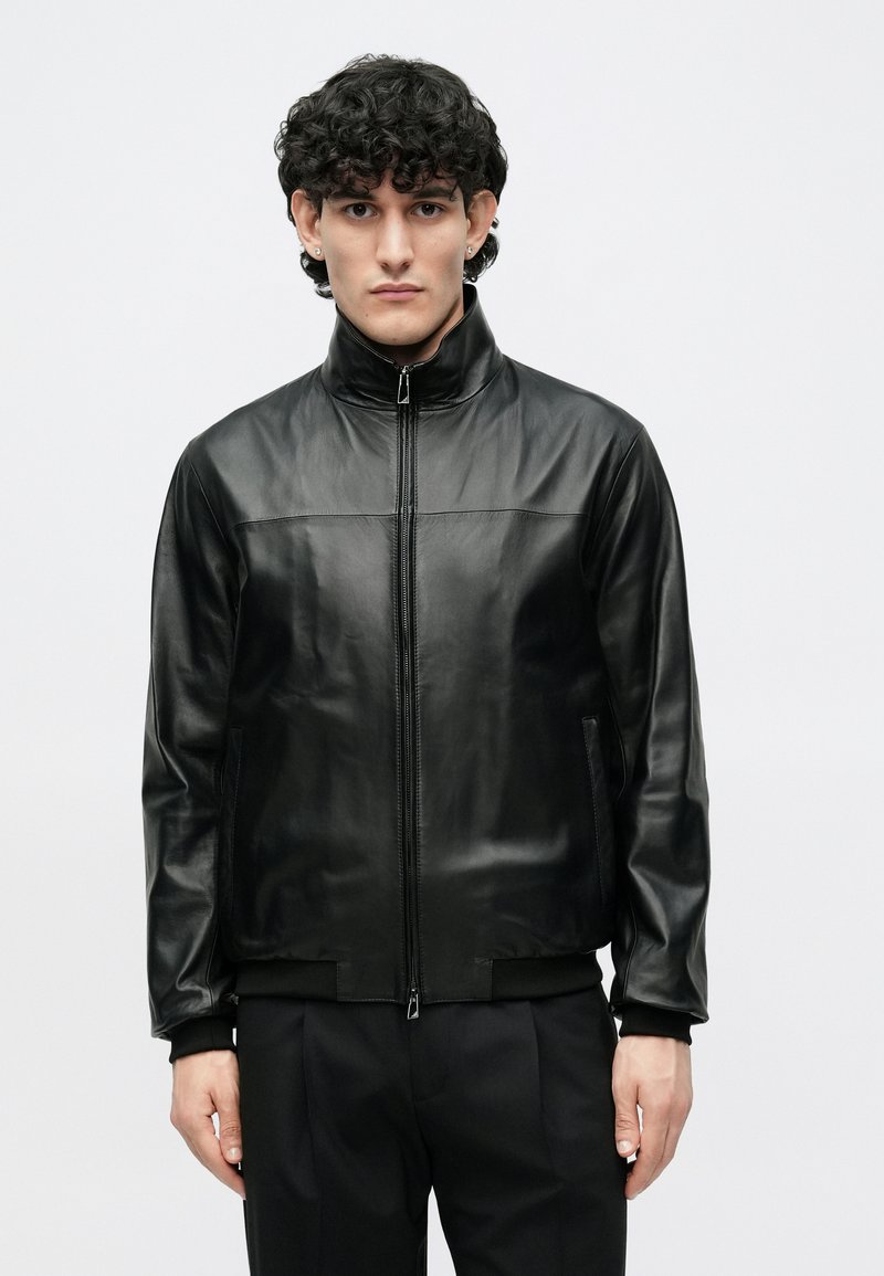 Veste en cuir noir avec col haut, fermeture éclair et poches latérales. Texture lisse et coupe ajustée, associée à un pantalon noir.