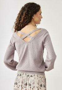 Suéter lila claro con un escote amplio y diseño de tirantes cruzados en la parte posterior. Material suave, mangas largas y cinturilla ajustada. Falda floral visible debajo.