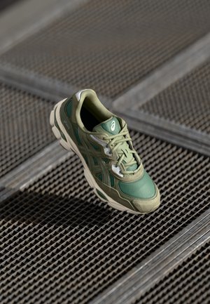Chaussure de sport verte et beige flottant au-dessus d'une surface en grille métallique à texture à motifs et encadrement argenté.