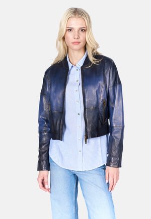 Veste en cuir bleu coupée avec une texture lisse, poches zippées et col montant, portée sur une chemise à rayures claire.