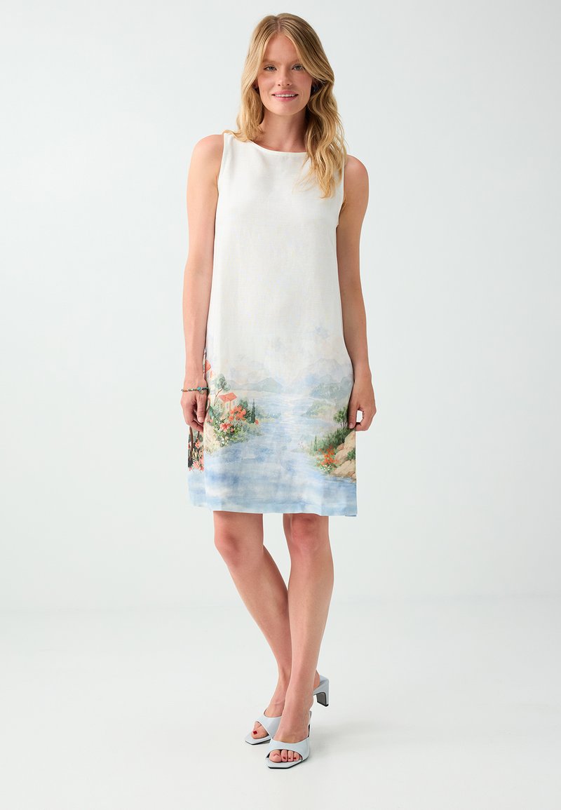 Femme debout portant une robe blanche sans manches avec un motif paysage en bas et des talons ouverts bleu clair sur un fond uni.