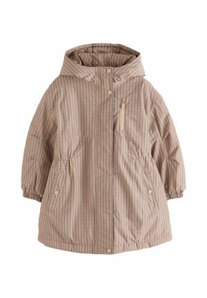 Lächelndes junges Mädchen mit geflochtenem Haar trägt einen beigen karierten Mantel, einen cremefarbenen Pullover und schwarze Stiefeletten vor einem einfarbigen Hintergrund.