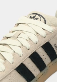 Beige mockasinsneaker med svart textilrand, krämfärgade snören och en rund logotyp på tungan. Märkbara vågiga detaljer på ränderna.