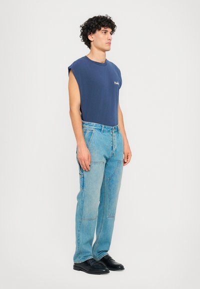 Marineblå ærmeløs top, lyseblå denimbukser, sorte sko. Bukserne har lommer og sømme; blødt bomuldsmateriale. Afslappet outfit design.