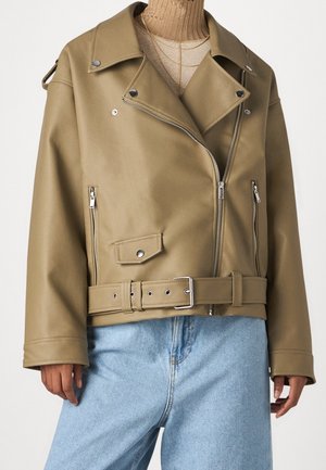 Veste biker kaki avec fermetures éclair argentées, boutons-pression, boucle de ceinture, portée sur un col roulé côtelé beige et un jean bleu clair.