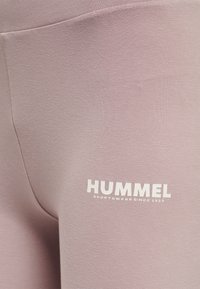 Leggings atletici rosa chiaro realizzati in un tessuto morbido; presentano un logo bianco con la scritta "HUMMEL SPORTSWEAR SINCE 1923" sulla coscia.