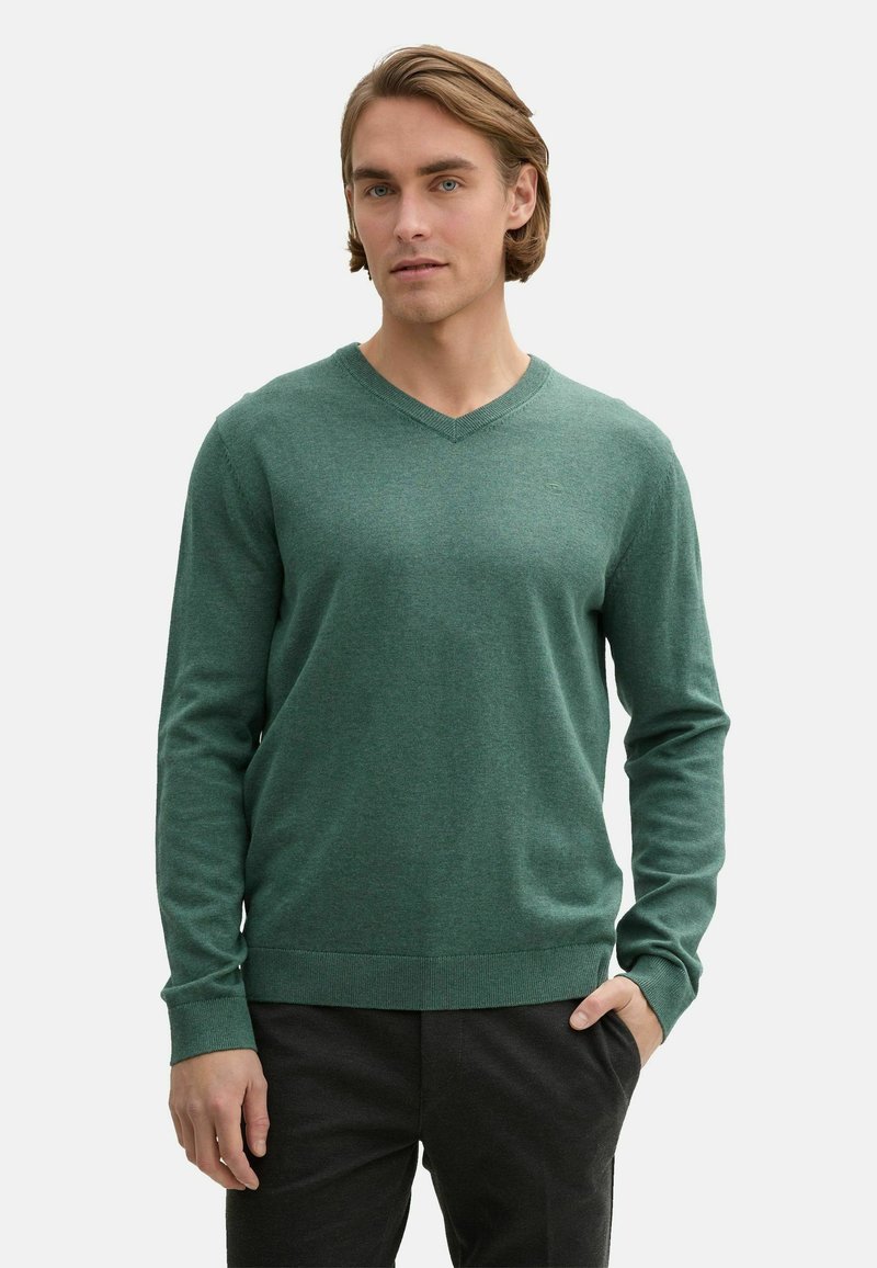 Pulover verde, cu mâneci lungi și decolteu în V, realizat dintr-un material tricotat moale. Are manșete și margine cu model din jarse. Este purtat cu pantaloni închiși la culoare, evidențiind o croială lejeră.