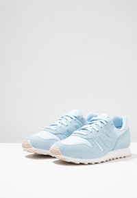 Baskets bleu clair avec des dessus en daim et en maille, accents blancs et lacets plats. Elles présentent une semelle texturée et un logo proéminent sur le côté.