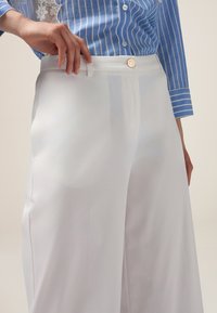 Pantalon large blanc avec une texture lisse, doté d'une fermeture à boutons à l'avant et de passants de ceinture, porté avec une chemise rayée bleue.