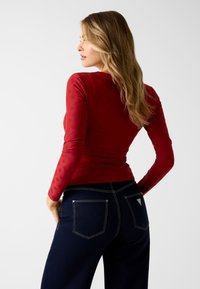 Rode top met lange mouwen en textuurpatroon, gecombineerd met donkerblauwe jeans met zichtbare naden en twee achterzakken, slanke pasvorm.