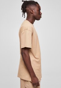 Tricou din bumbac bej, cu mâneci scurte, croială lejeră, decolteu rotund și tiv drept. Profilul lateral al modelului evidențiază tăietura și structura articolului.