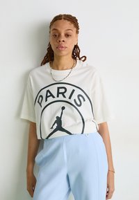 T-shirt blanc surdimensionné avec un graphique noir "PARIS" et une silhouette d'un joueur. Associé à un pantalon ajusté bleu clair.