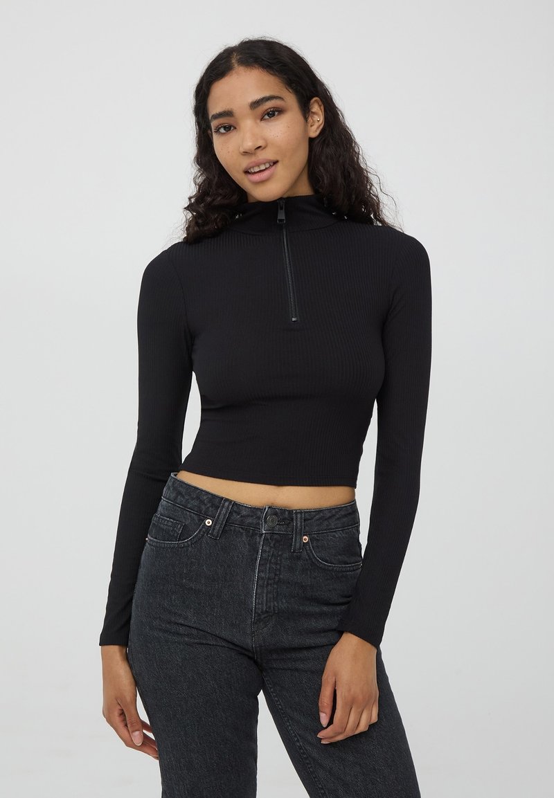 Maglietta nera a maniche lunghe e croppata con colletto alto e chiusura a mezza cerniera, realizzata in tessuto a costine. Abbinata a jeans scuri.