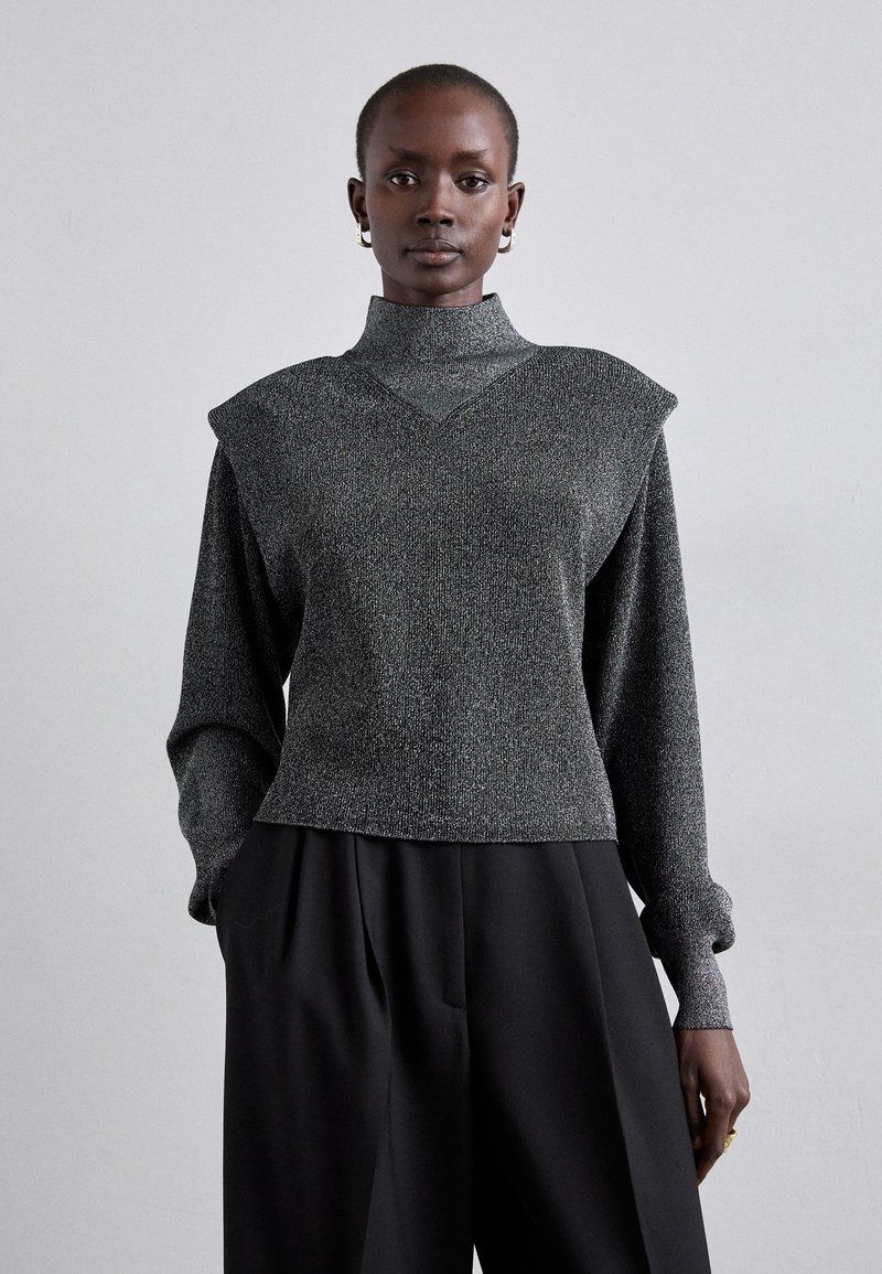 Top gris con textura, cuello alto, acentos en los hombros y mangas largas; combinado con pantalones negros de corte holgado.