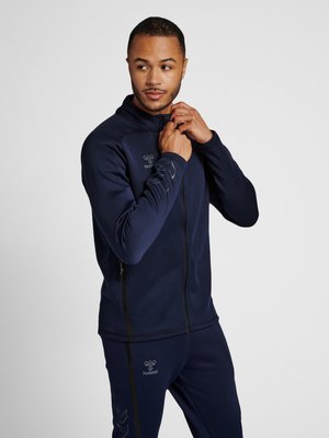 Man die een marineblauwe sportjas met bijpassende broek dichtritst tegen een effen witte achtergrond.