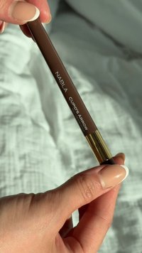 Crayon Nabla Cupid's Arrow, corps brun avec des accents dorés, doté d'une pointe fine. Main tenant le crayon contre un fond texturé clair.