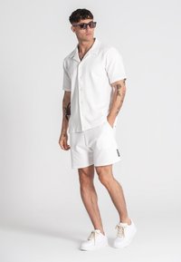 Camisa blanca de manga corta con botones y pantalones cortos texturizados a juego. El calzado son zapatillas blancas con cordones crema. Los accesorios incluyen gafas de sol.