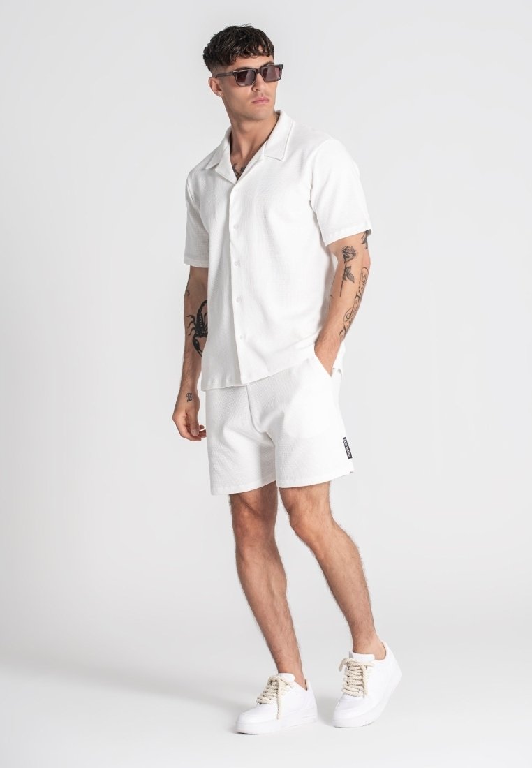 Camisa blanca de manga corta con botones y pantalones cortos texturizados a juego. El calzado son zapatillas blancas con cordones crema. Los accesorios incluyen gafas de sol.
