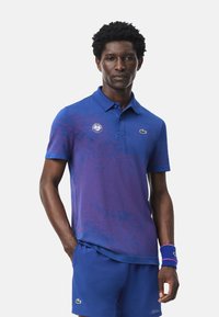 Blå och lila gradientpolo med krage, som har en logo på bröstet och en subtil textur. Matchas med enfärgade blå shorts.