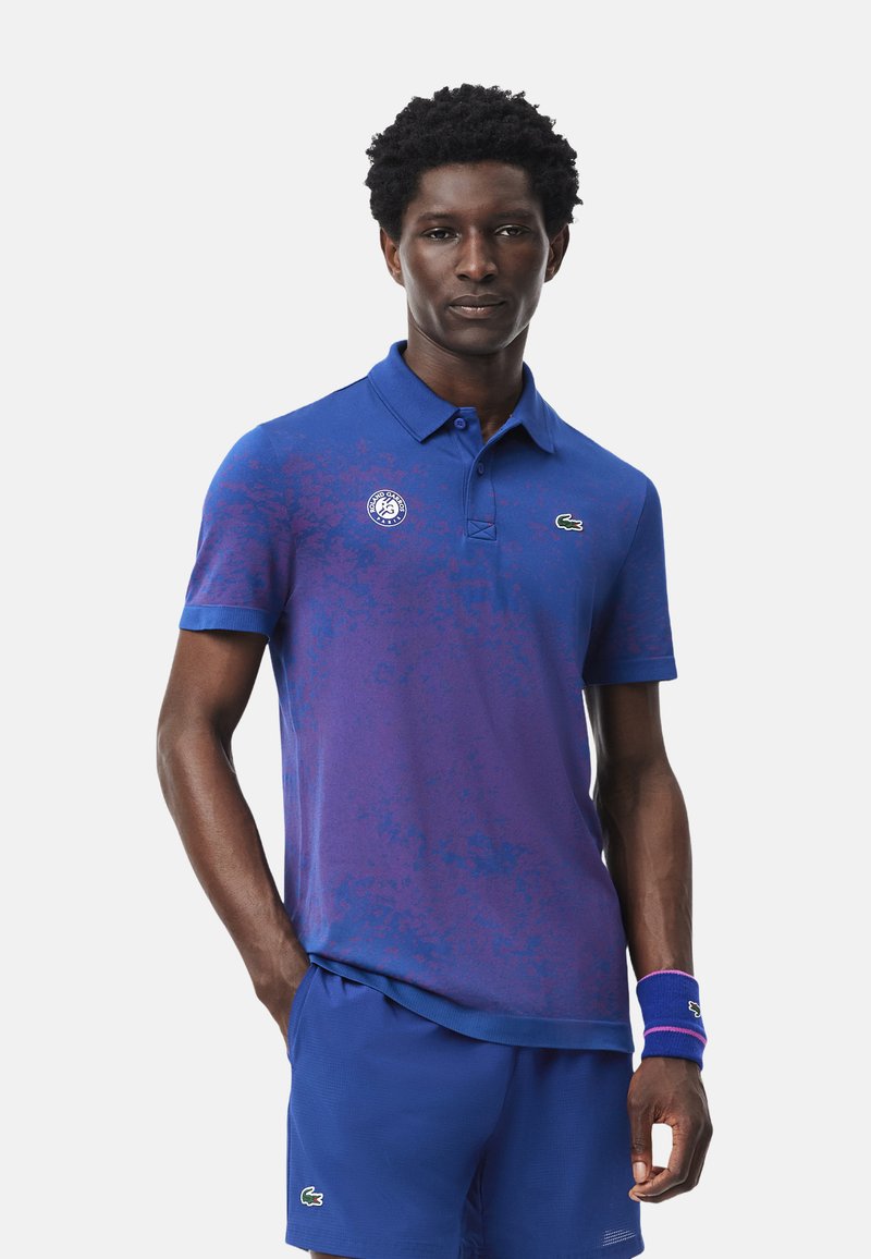 Blå och lila gradientpolo med krage, som har en logo på bröstet och en subtil textur. Matchas med enfärgade blå shorts.