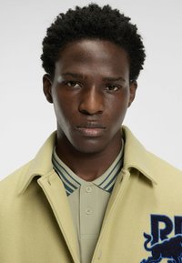 Kremowa wełniana kurtka z kołnierzem, ozdobiona haftowanym niebieskim logo lwa. Nałożona na jasnoszary polo z paskowymi akcentami.