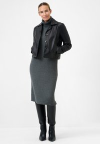 Schwarze, kurz geschnittene Lederjacke mit silbernen Knöpfen, über einem grauen, gerippten Strickkleid getragen. Das Modell trägt schwarze, knielange Stiefeletten mit Absatz.