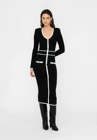 Zwarte gebreide jurk met een diepe V-hals, witte knoopsluiting en zakken, met zilverkleurige spikkels en lange mouwen. Gestyled met kniehoge laarzen.
