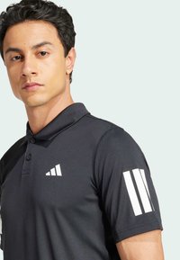 Czarna koszulka polo wykonana z gładkiego materiału, z kołnierzykiem, trzema białymi paskami na rękawie i białym logo na klatce piersiowej.