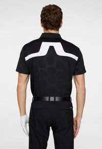 Black polo shirt with hexagonal pattern, featuring a white geometric design on the upper back; paired with black pants and a white golf glove.