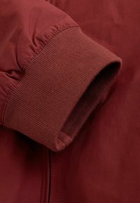 Poignet de veste bordeaux avec tricot côtelé élastique, tissu lisse à finition mate, et une couture dissimulée le long du bord.