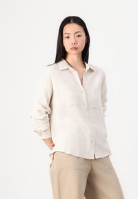 Lichtbeige overhemd met knoopsluiting en een kraag, twee borstzakken en opgerolde mouwen, gemaakt van gestructureerde stof. G gedragen met beige broek.