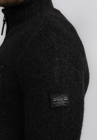 Zwarte gebreide trui met hoge kraag, voorzien van een ritssluiting en een rechthoekig label op de linkerarm met witte tekst.