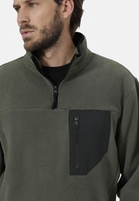 Olivgrüner Fleece-Pullover mit einem Viertel-Reißverschluss-Kragen, ausgestattet mit einer schwarzen, mit Reißverschluss versehenen Brusttasche und einem weichen, strukturierten Stoff.