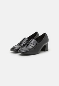 Chaussures noires en cuir verni avec un petit talon carré, présentant une pointe arrondie et des accents de sangle décoratifs sur le cou-de-pied.