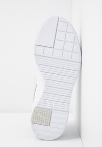 Semelle de sneaker blanche en caoutchouc texturé, avec un motif en grille et un détail de logo emblématique. Le profil latéral montre un design de talon arrondi.