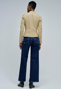 Femme portant une veste en cuir beige, un jean large bleu foncé et des bottes à talons noires, se tenant dos à un fond uni.