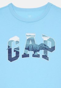 Világoskék, rövid ujjú póló, amelyen egy nagy "GAP" logó található, a betűkben dinoszaurusz grafikával, textúrázott háttér előtt.