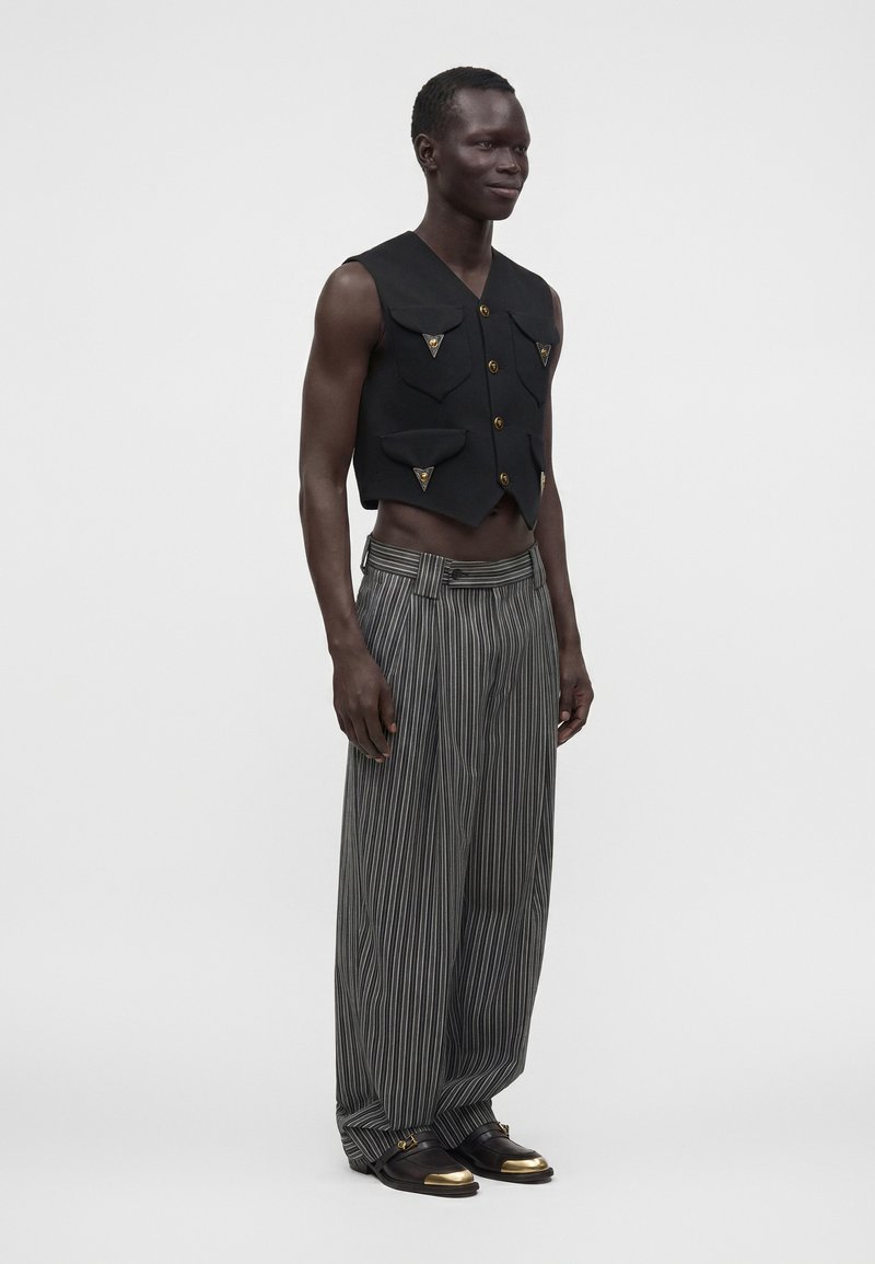 Homme debout portant un gilet noir sans manches avec poches, un pantalon ample gris à fines rayures et des chaussures noires avec embouts métalliques dorés.