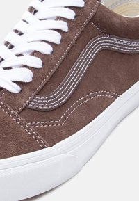 Bruine suède sneaker met witte veters, witte rubberen zool en een kenmerkend wit stikselpatroon op het zijpaneel.