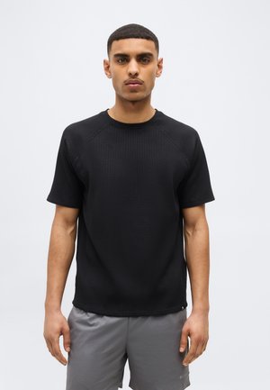 Fiatal férfi rövid hajjal, fekete texturált rövid ujjú ingben és szürke rövidnadrágban, egyszínű világos háttér előtt.