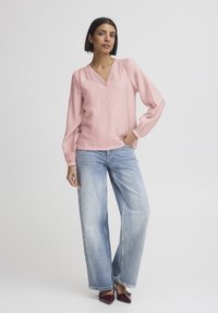 Světle růžová halenka s dlouhými rukávy, výstřihem do V a nařasenými rameny, ve spojení se světle modrými džíny s širokými nohavicemi a vínově červenými botami s špičkou.