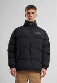 Jachetă puffer neagră cu guler înalt, închidere cu fermoar și manșete elastice. Are un quilt vertical și logo-ul "KANI" pe piept.