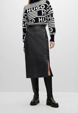 Kobieta ubrana w czarno-biały wzorzysty sweter HUGO, czarną skórzaną spódnicę midi z wysokim stanem i bocznym rozcięciem oraz czarne kozaki za kolano.