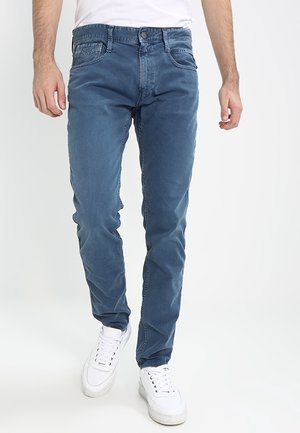 Man draagt strakke blauwe spijkerbroek, witte sneakers en een wit t-shirt, staand tegen een effen lichte achtergrond.